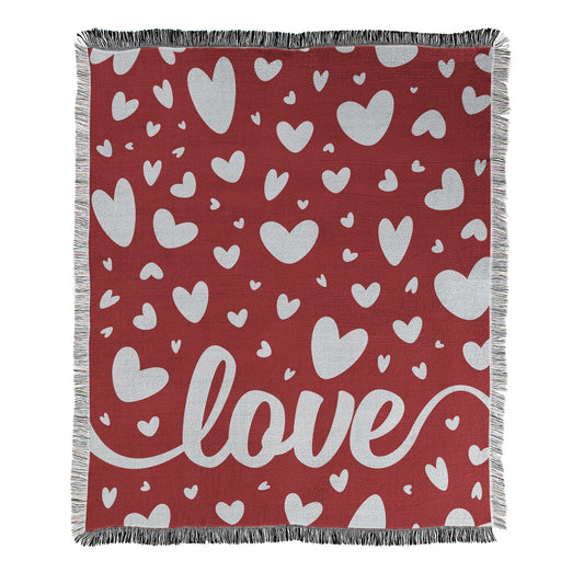 Red blanket with white hearts pattern showing a script text 'love' at the bottom, printed on a woven blanket with decorative fringed edges.