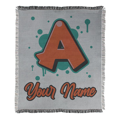 Graffiti style design printed with a letter and example text 'your name' on a blanket with decorative fringe edges.